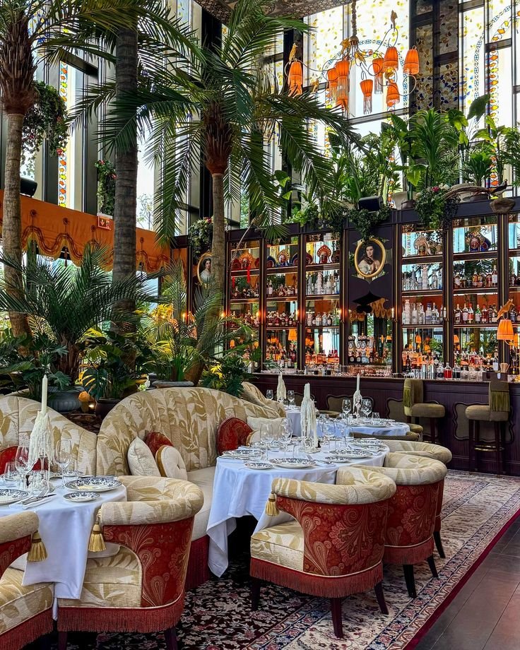 This may contain: a restaurant with many tables and chairs in front of large windows, decorated with palm trees