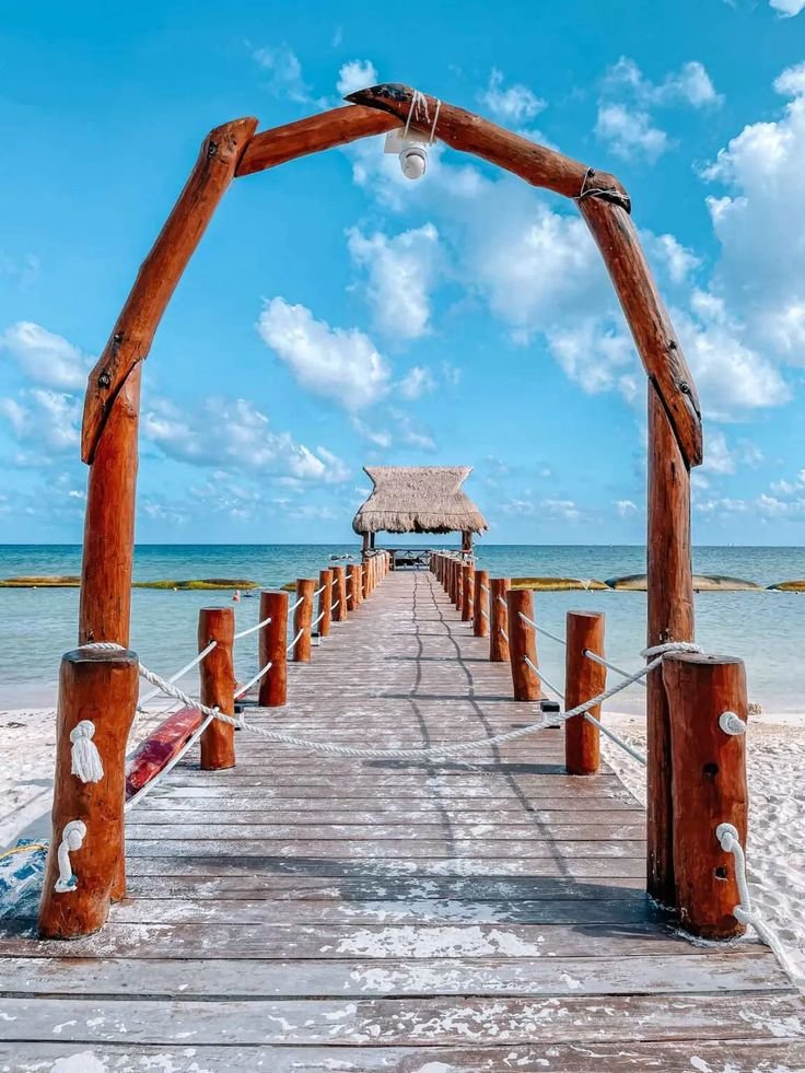 This may contain: a wooden pier on the beach with a thatched hut in the background and blue sky
