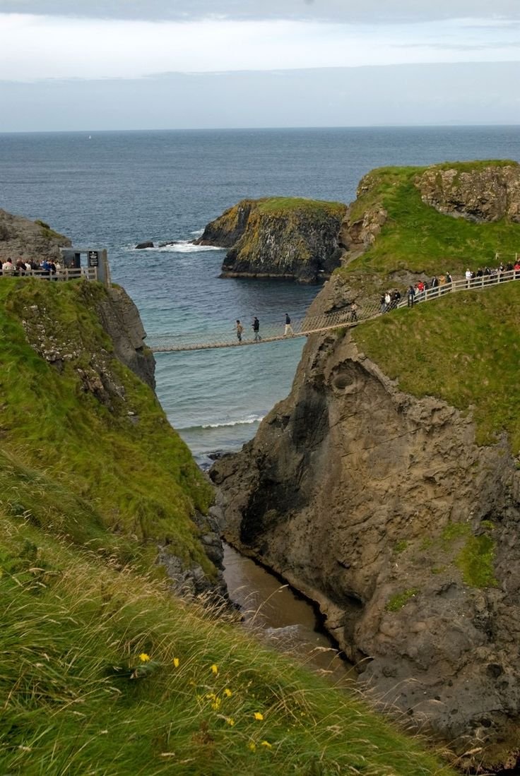 This may contain: people are walking on a bridge over the water near some rocks and grass, with cliffs in the background
