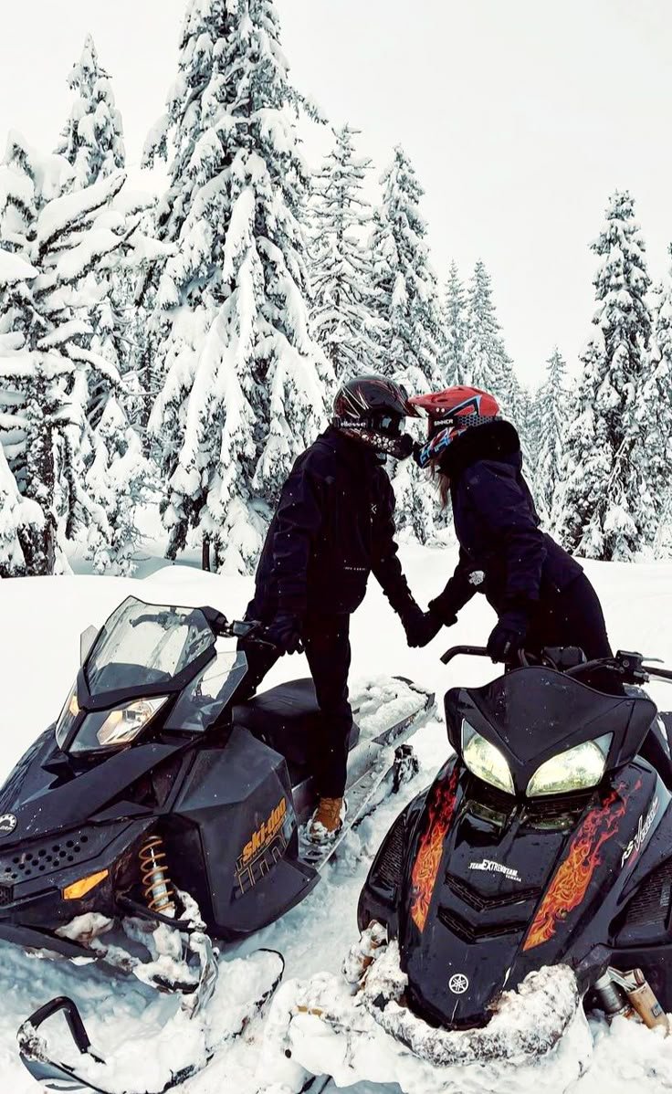 This may contain: two people on snowmobiles touching hands in the snow with trees in the background