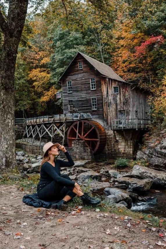 This may contain: a woman sitting on the ground in front of a water mill with her hat on