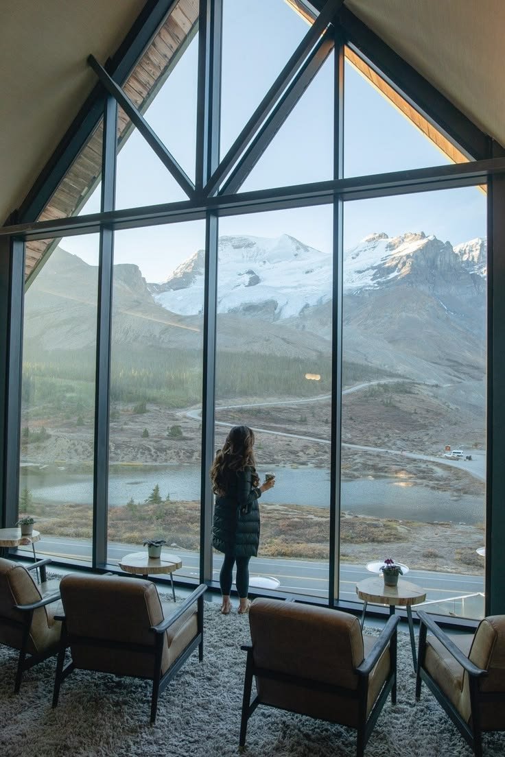 This may contain: a woman standing in front of a large window looking out at the mountains and lake