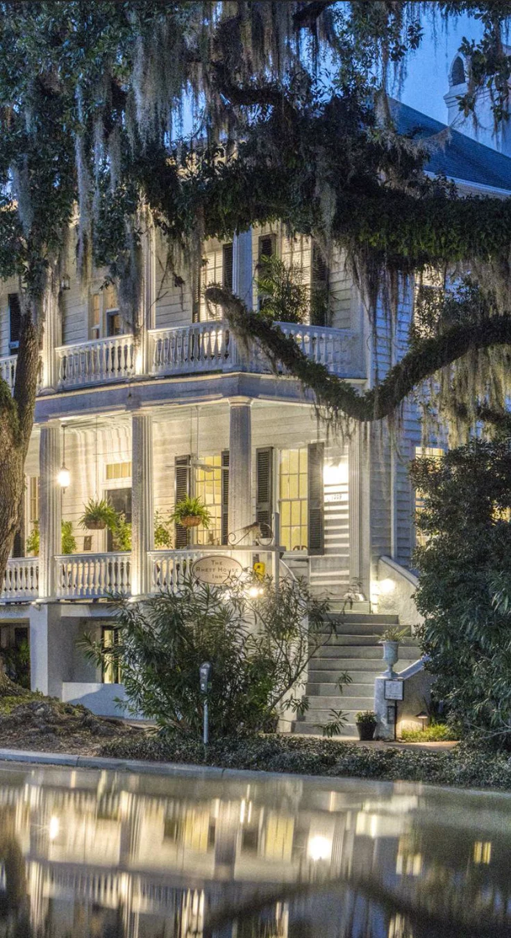 This may contain: a large white house sitting next to a body of water with trees in front of it