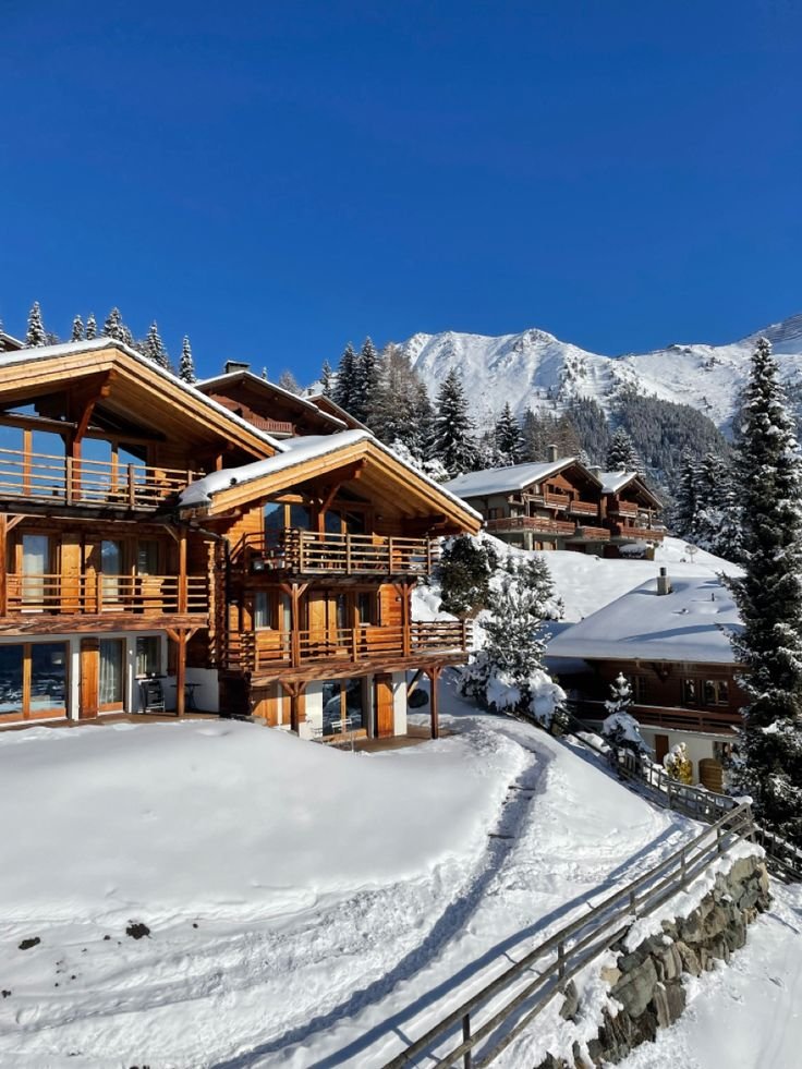 This may contain: a large wooden house on top of a snow covered slope with mountains in the background