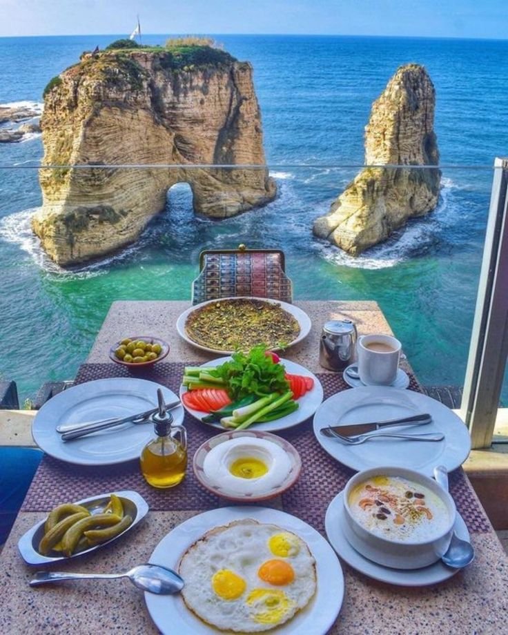 This may contain: an outdoor dining table with food and drinks on it overlooking the ocean in front of two rock formations