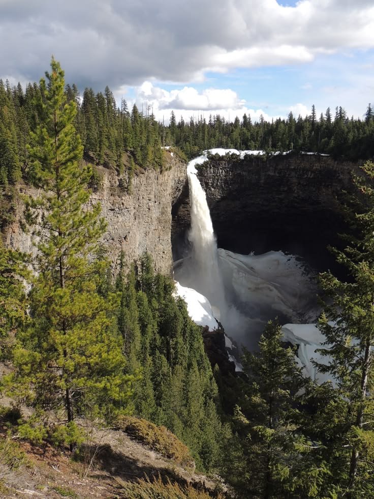This may contain: a waterfall with snow on the ground and trees around it, surrounded by evergreens