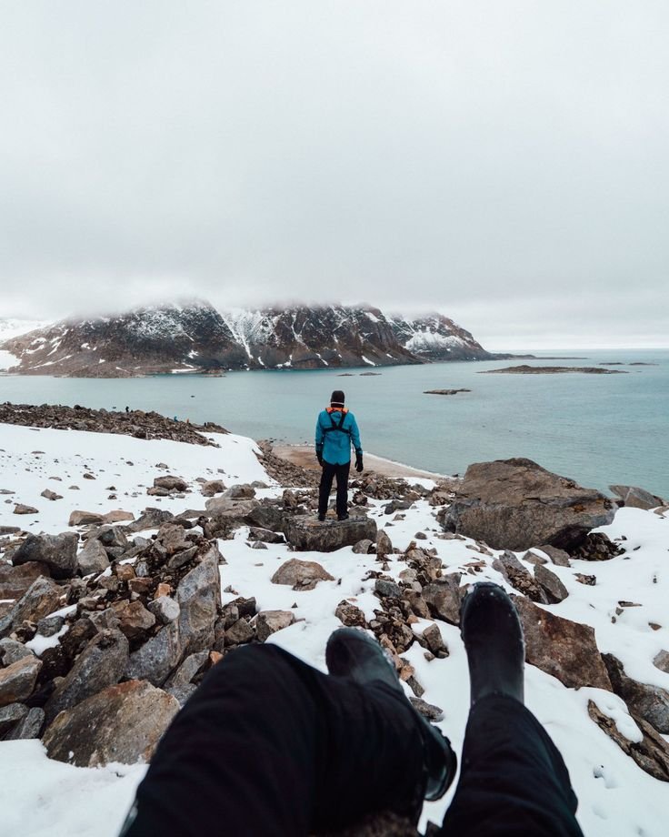 This may contain: a person standing on top of a snow covered hill next to the ocean with their feet in the air