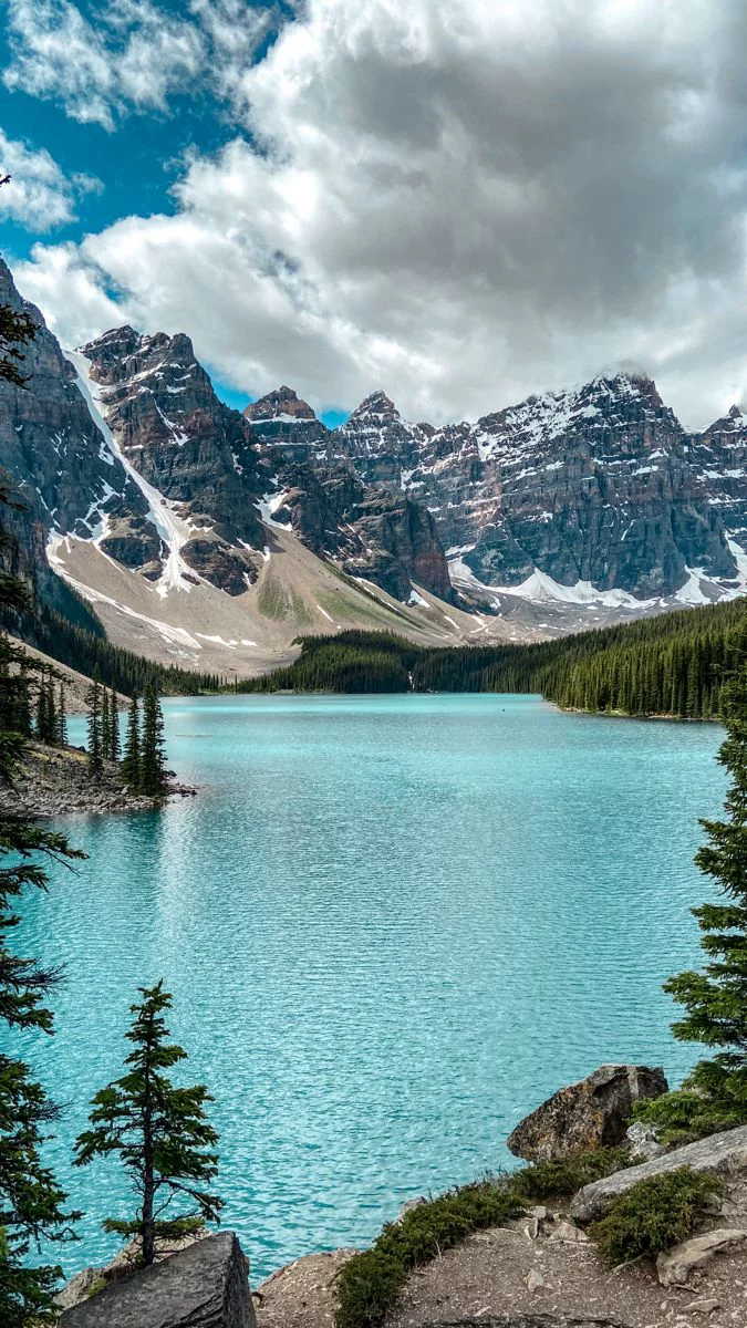 This may contain: a lake surrounded by mountains and trees under a cloudy sky