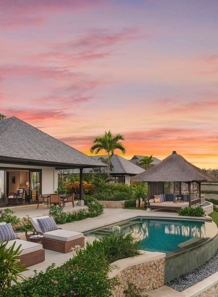 This may contain: an outdoor pool with lounge chairs and thatched umbrellas in the background at sunset