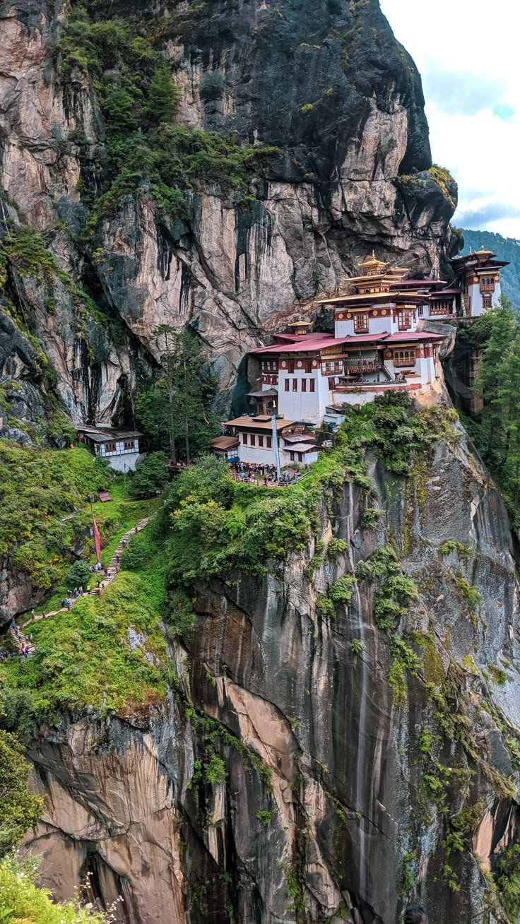 This may contain: a mountain side with some buildings on top of it and trees growing out of the cliff