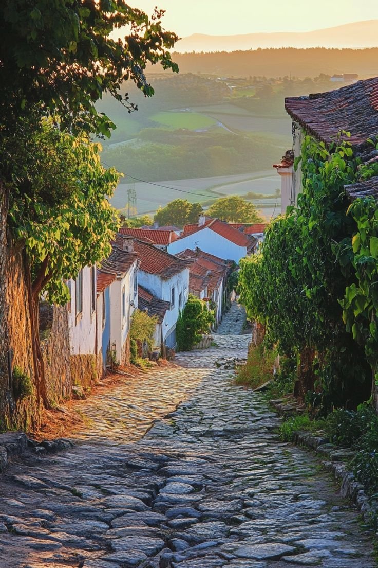 This may contain: an old cobblestone street with houses and hills in the background at sunset or sunrise