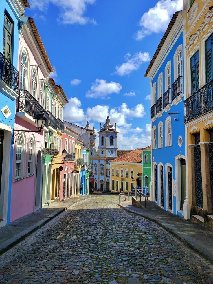 This may contain: a cobblestone street lined with brightly colored buildings under a blue sky filled with clouds
