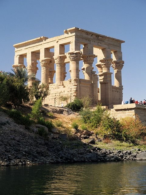 This may contain: an old building sitting on top of a hill next to a body of water with people standing around it