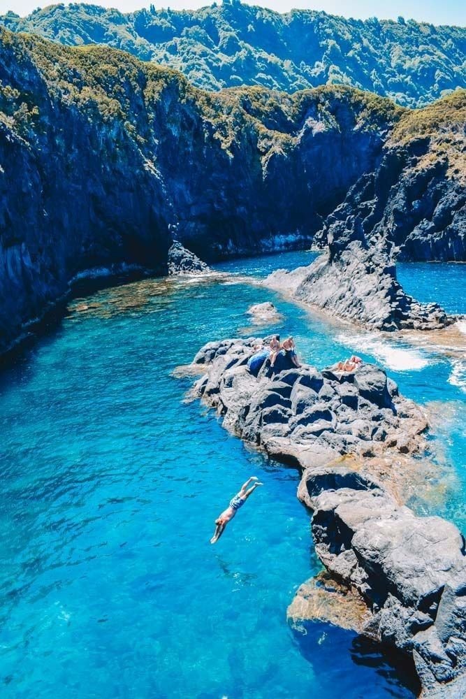 This may contain: people are swimming in the blue water near some rocks and mountains with green trees on either side