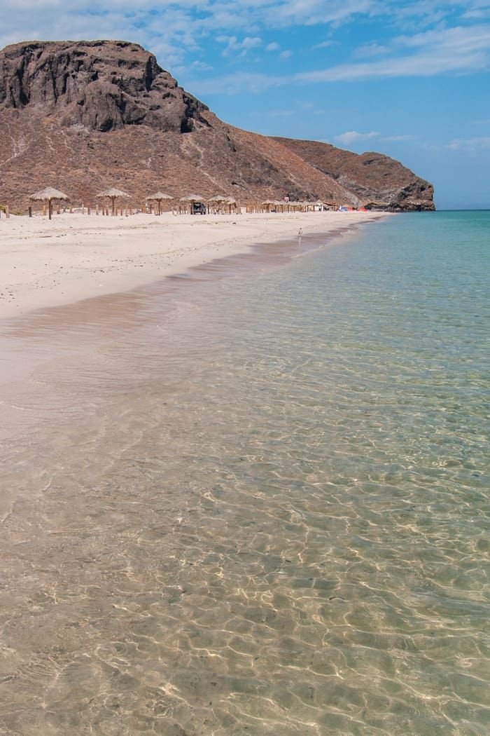 This may contain: an empty beach with clear water and mountains in the background