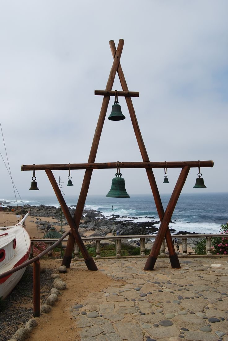 This may contain: a wooden structure with bells hanging from it's sides next to the ocean and a boat