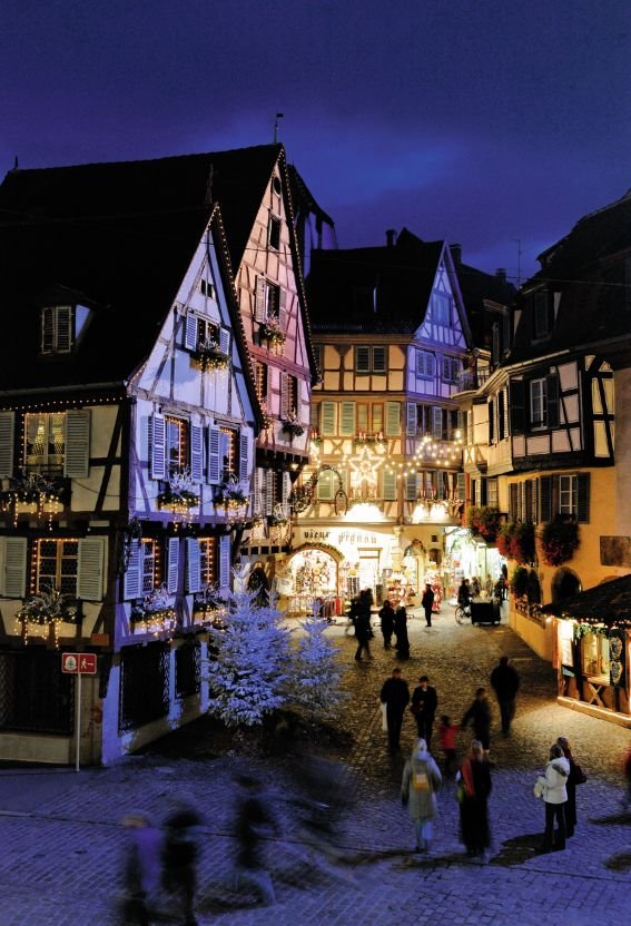 This may contain: people are walking around in an old european town at christmas time with lights and decorations on the buildings