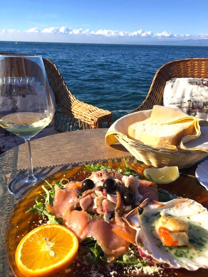 This may contain: a plate of food on a table next to the ocean with two glasses of wine