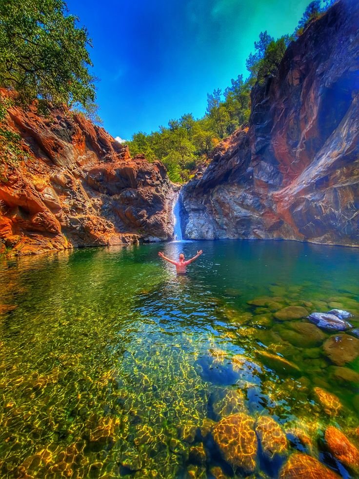 This may contain: a person swimming in a body of water surrounded by rocks and trees, under a blue sky