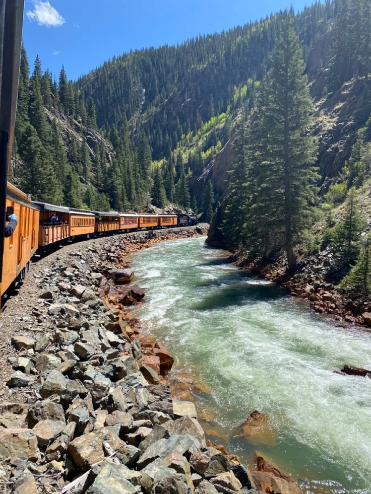 This may contain: a train traveling down tracks next to a river with rocks and trees on both sides