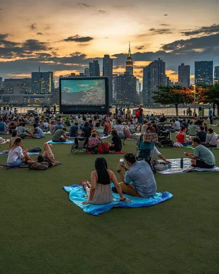 This may contain: many people are sitting on the grass in front of an outdoor movie screen at sunset