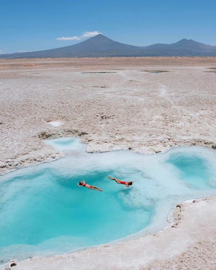 This may contain: two people are swimming in a pool of blue water with mountains in the back ground