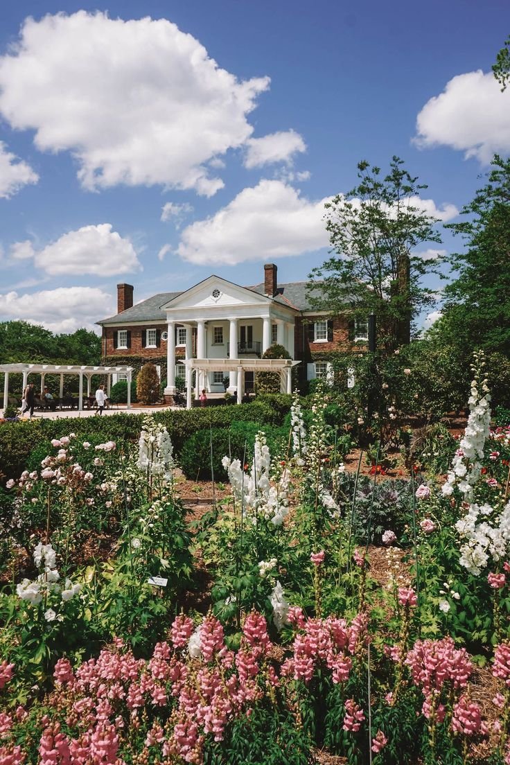 This may contain: a large house surrounded by lots of flowers and greenery on a sunny day with clouds in the sky