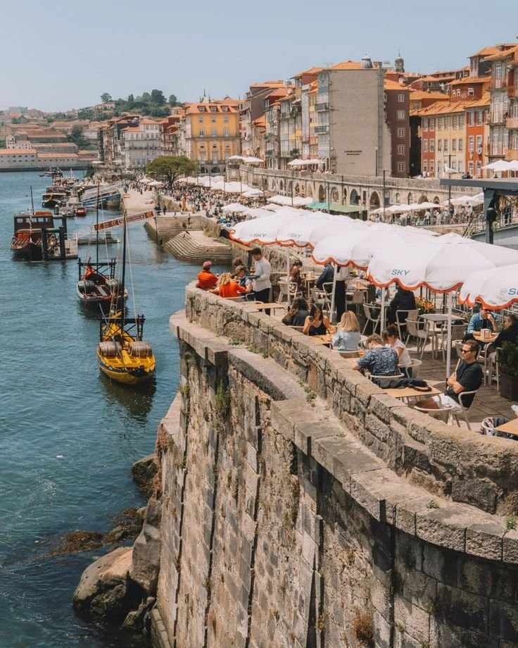 This may contain: people are sitting at tables on the edge of a cliff by the water with boats in the background