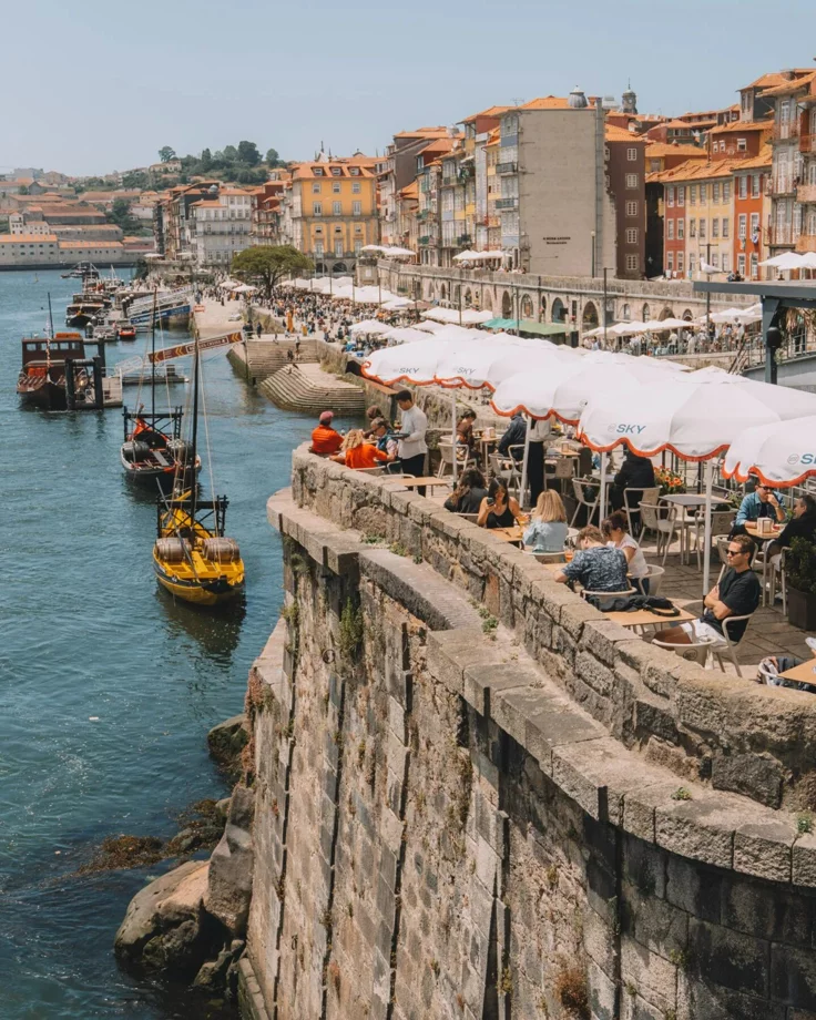This may contain: people are sitting at tables on the edge of a cliff by the water with boats in the background