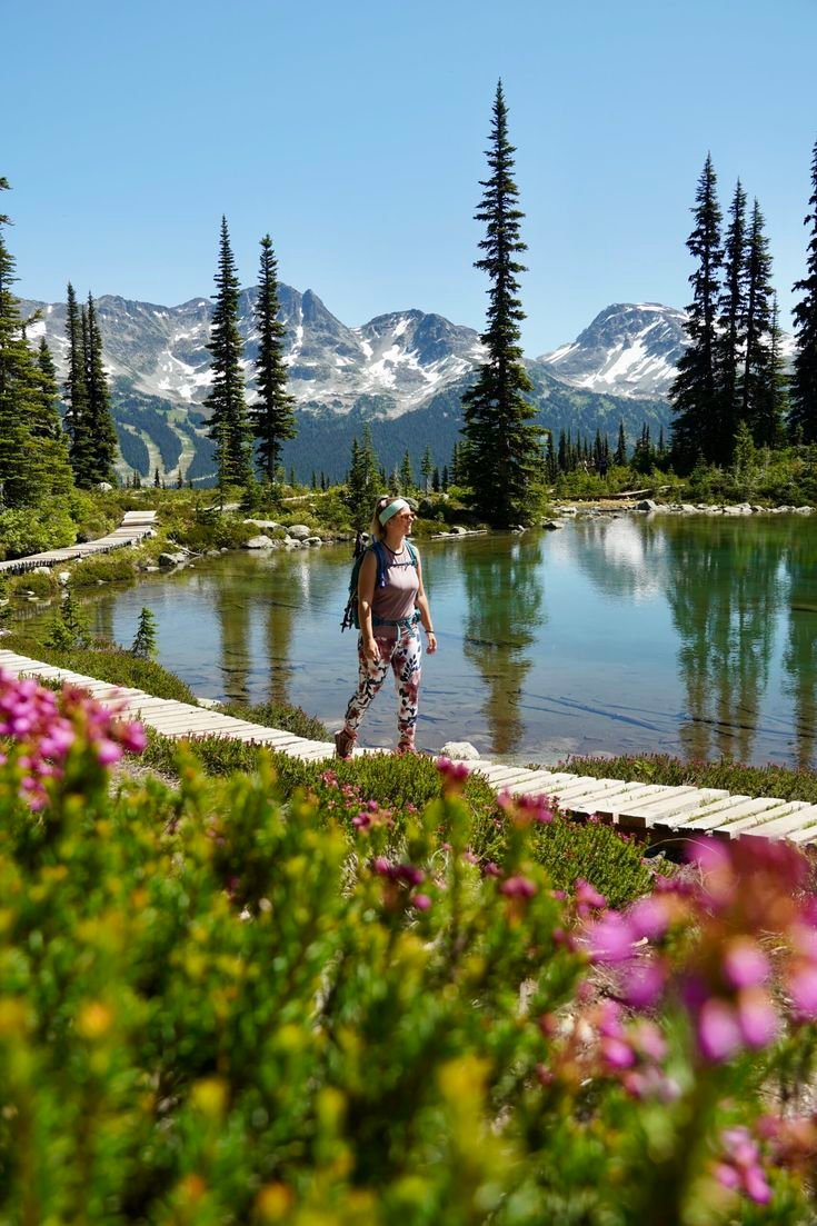 This may contain: a woman walking across a lush green field next to a lake with mountains in the background