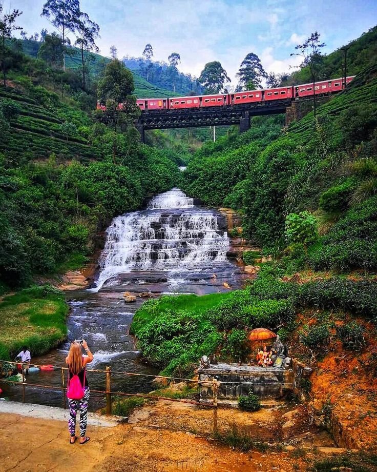 This may contain: a woman standing in front of a waterfall with a train on the track above it
