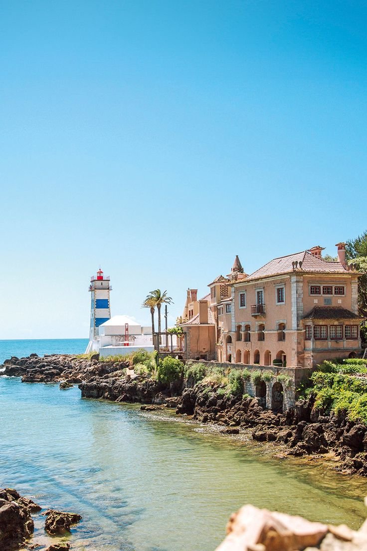 This may contain: the lighthouse is next to some houses on the shore by the water with palm trees in the foreground