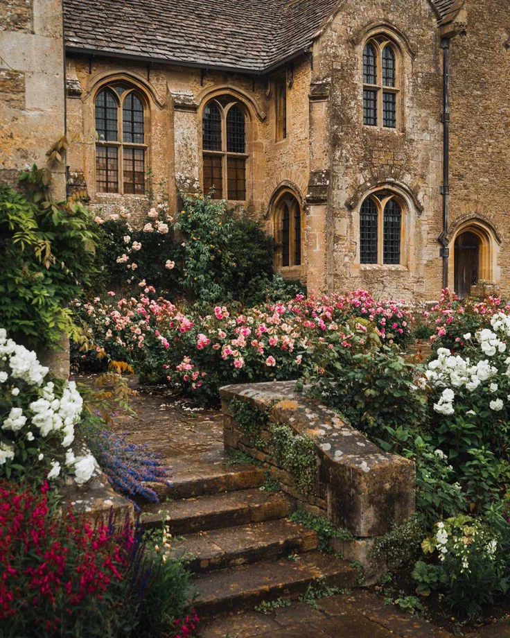 This may contain: an old stone house with flowers in the foreground and steps leading up to it