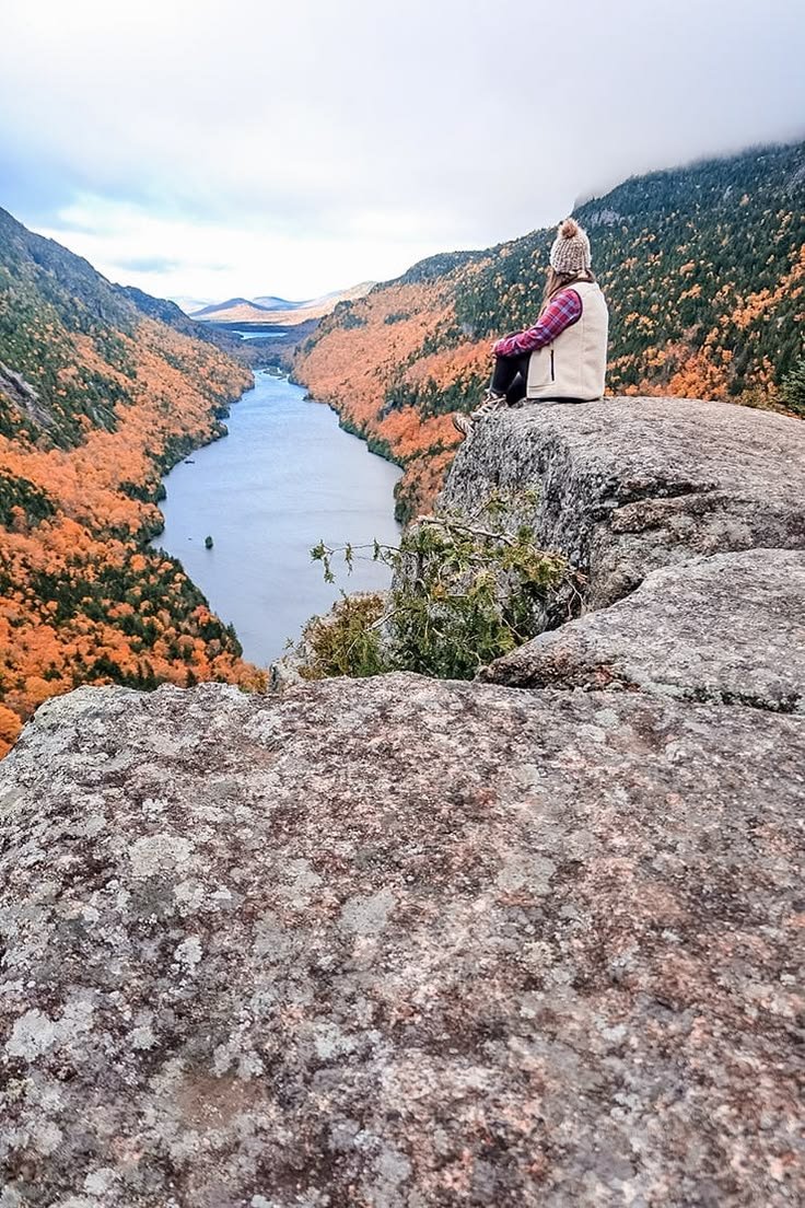 This may contain: a woman sitting on top of a large rock next to a lake in the mountains