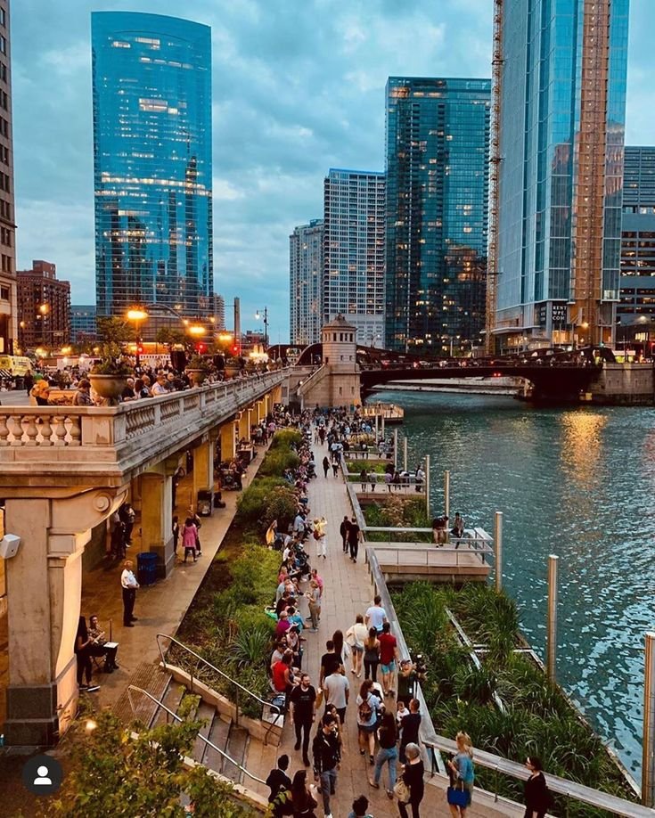 This may contain: many people are walking on the sidewalk by the water and buildings in the city at night