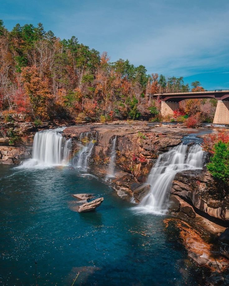 This may contain: a large waterfall in the middle of a river with a bridge above it and trees surrounding it
