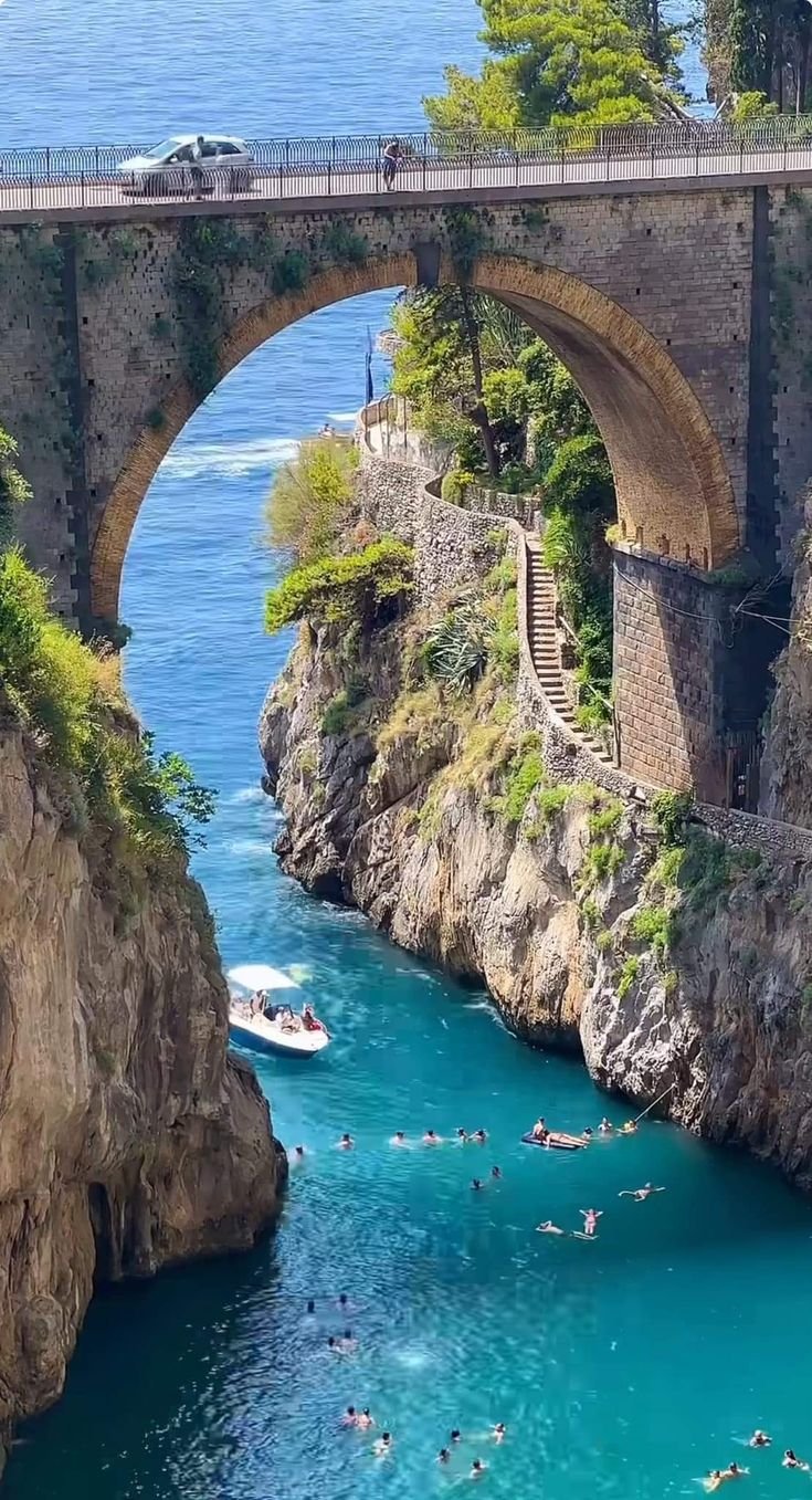 This may contain: people are swimming in the blue water under an old bridge that is built on top of a cliff