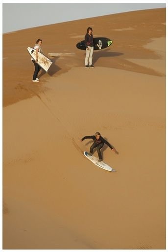 This may contain: three surfers carrying their boards across the sand dunes in the desert, one is falling off his surfboard