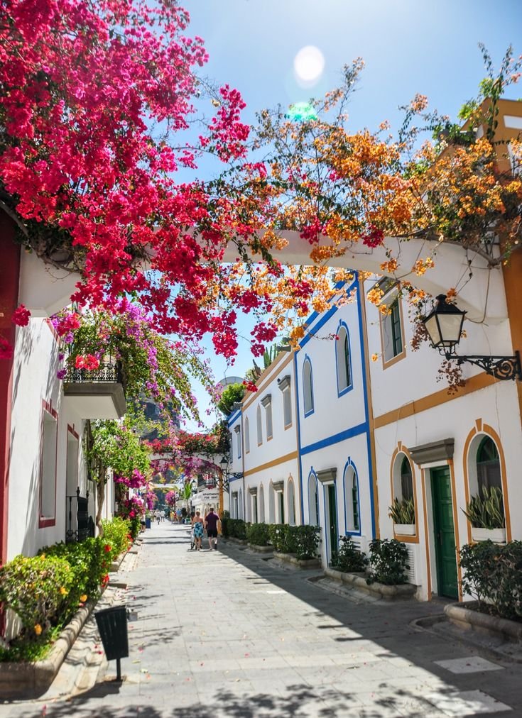 This may contain: a narrow street lined with white buildings and colorful flowers hanging from the ceiling over it