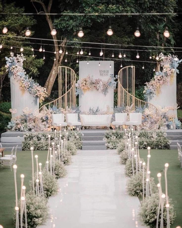 This may contain: an outdoor wedding ceremony setup with candles and floral arrangements on the aisle, surrounded by greenery