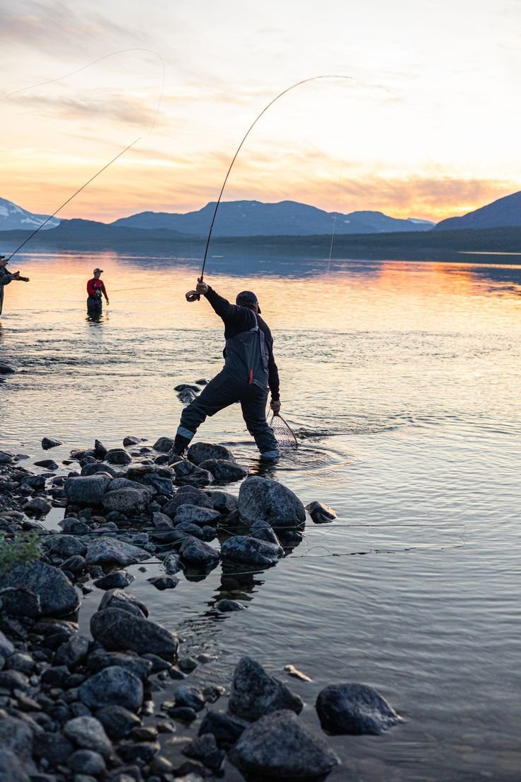 This may contain: two men are fishing in the water with rocks on the shore and mountains in the background