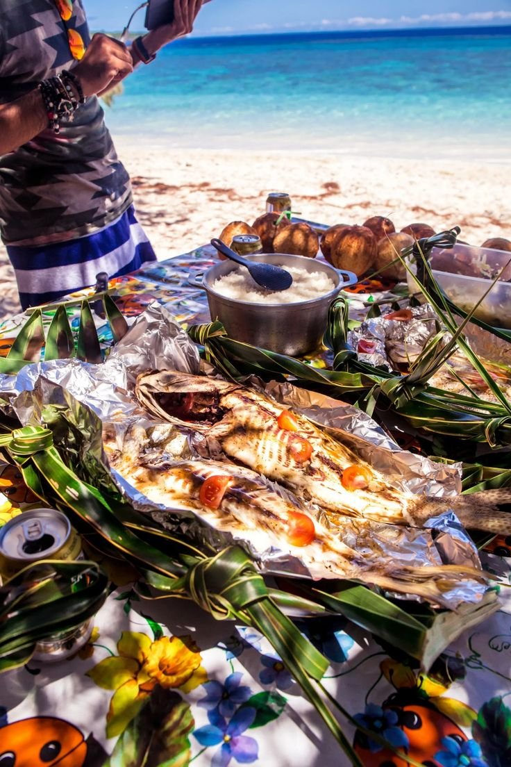This may contain: a man standing next to a table filled with food on top of a sandy beach