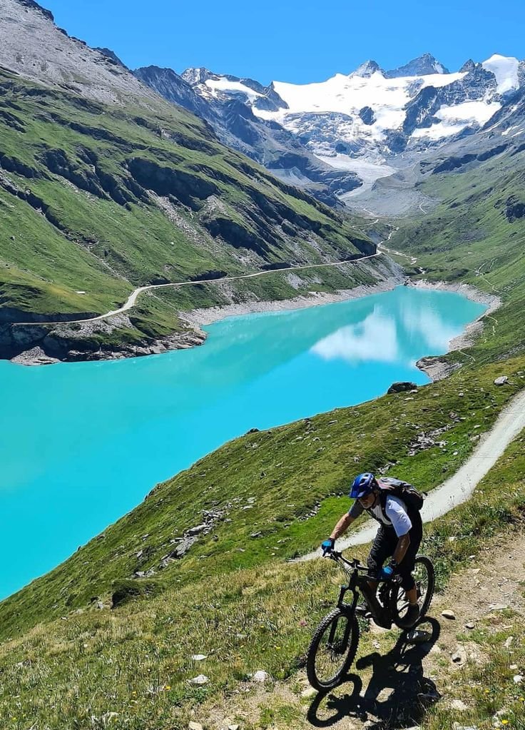 This may contain: a person riding a bike on a trail near a lake and mountains in the background