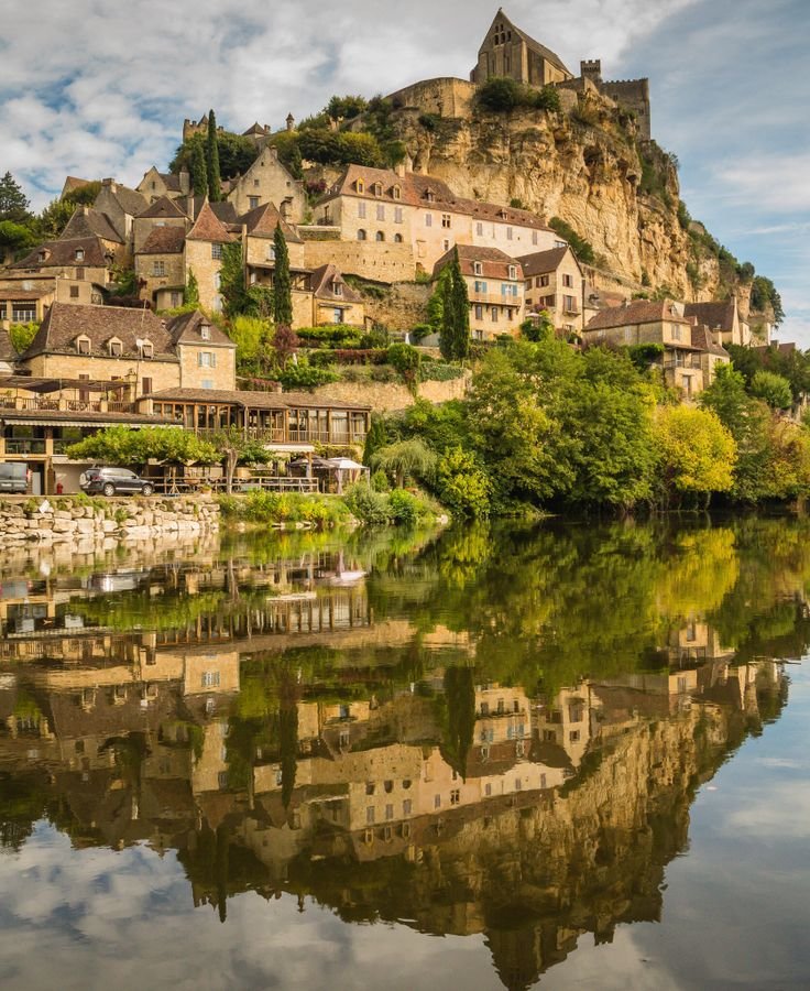 This may contain: an old town sits on top of a hill next to a river with reflections in the water