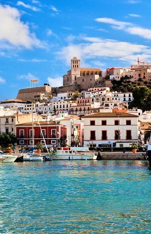 This may contain: boats are in the water near buildings on a hill behind them and blue sky with wispy clouds