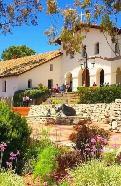 This may contain: an old church is surrounded by flowers and greenery on a sunny day with people walking around