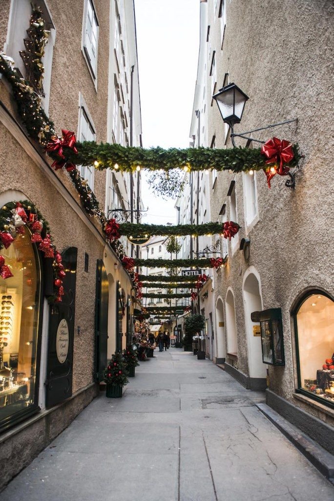This may contain: an alley way with christmas decorations and lights on the buildings along it's sides