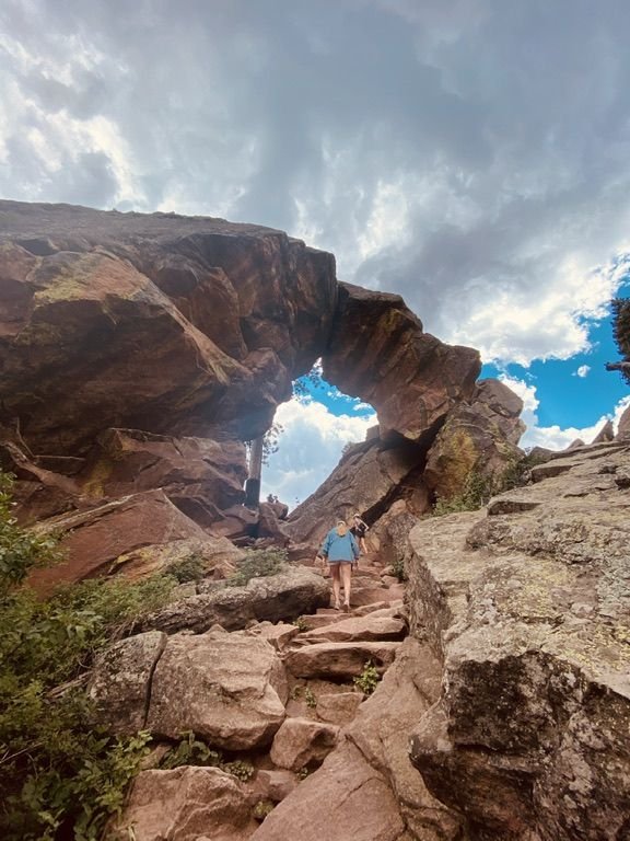 This may contain: a person is walking up some rocks on a trail with an arch in the background