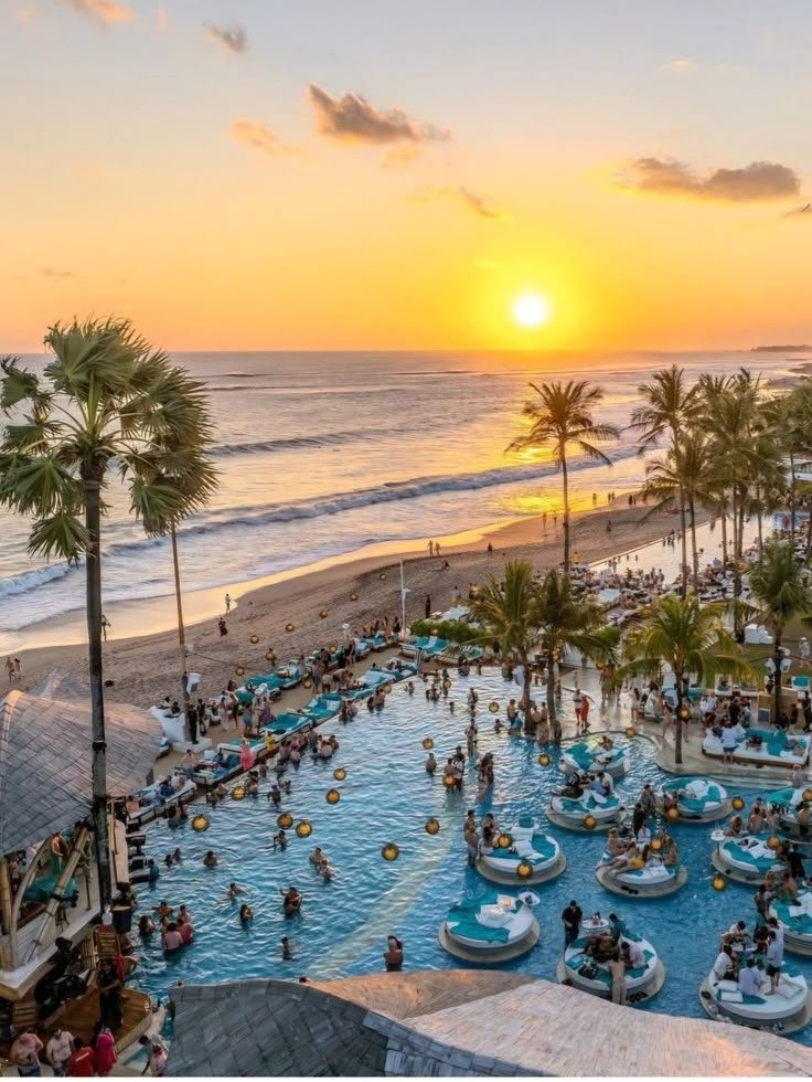 This may contain: the sun is setting over an outdoor pool with people swimming in it and palm trees lining the beach