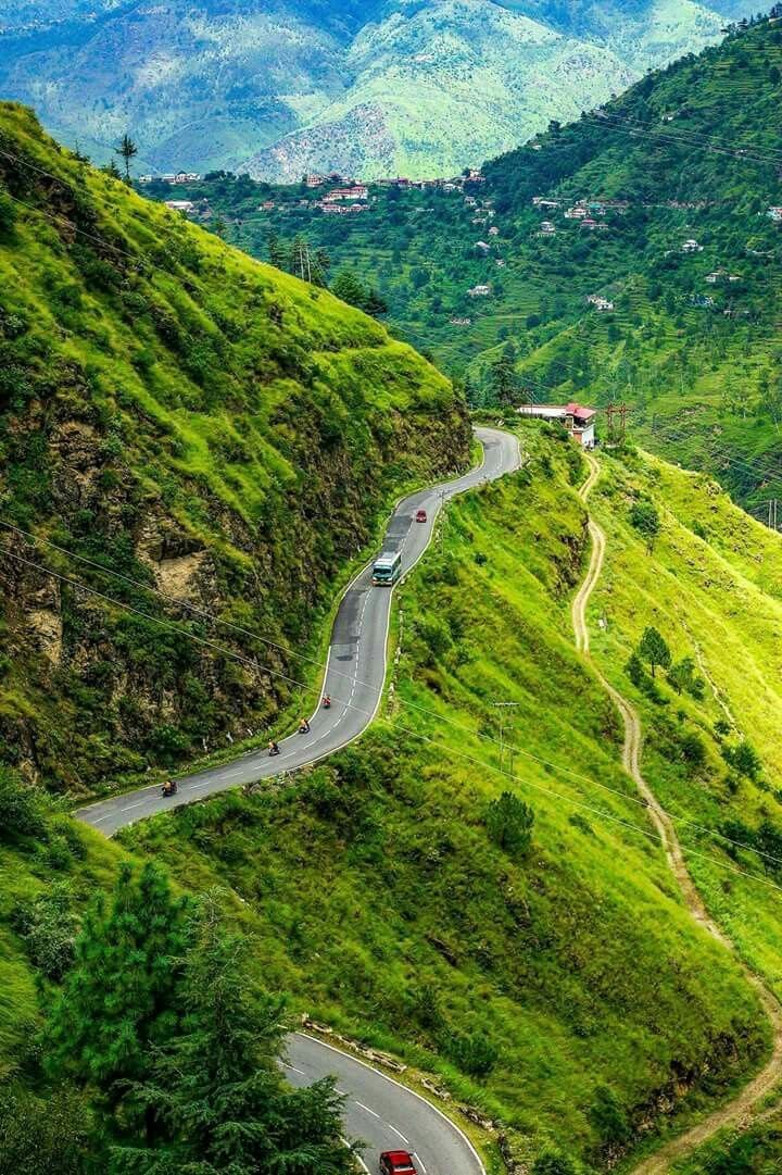 This may contain: a car driving down a winding road on top of a lush green hill side with mountains in the background